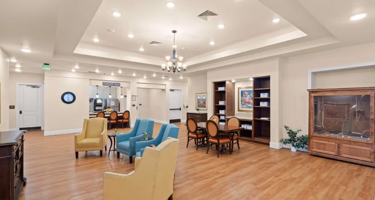A spacious, well-lit common area in a senior living facility featuring a seating arrangement with two yellow armchairs and two blue armchairs around small tables with flowers. There is a wooden table with four chairs near built-in shelves containing storage boxes and a framed picture. In the background, a coffee bar area with three chairs is visible. The room has wood flooring, recessed lighting, and a chandelier.