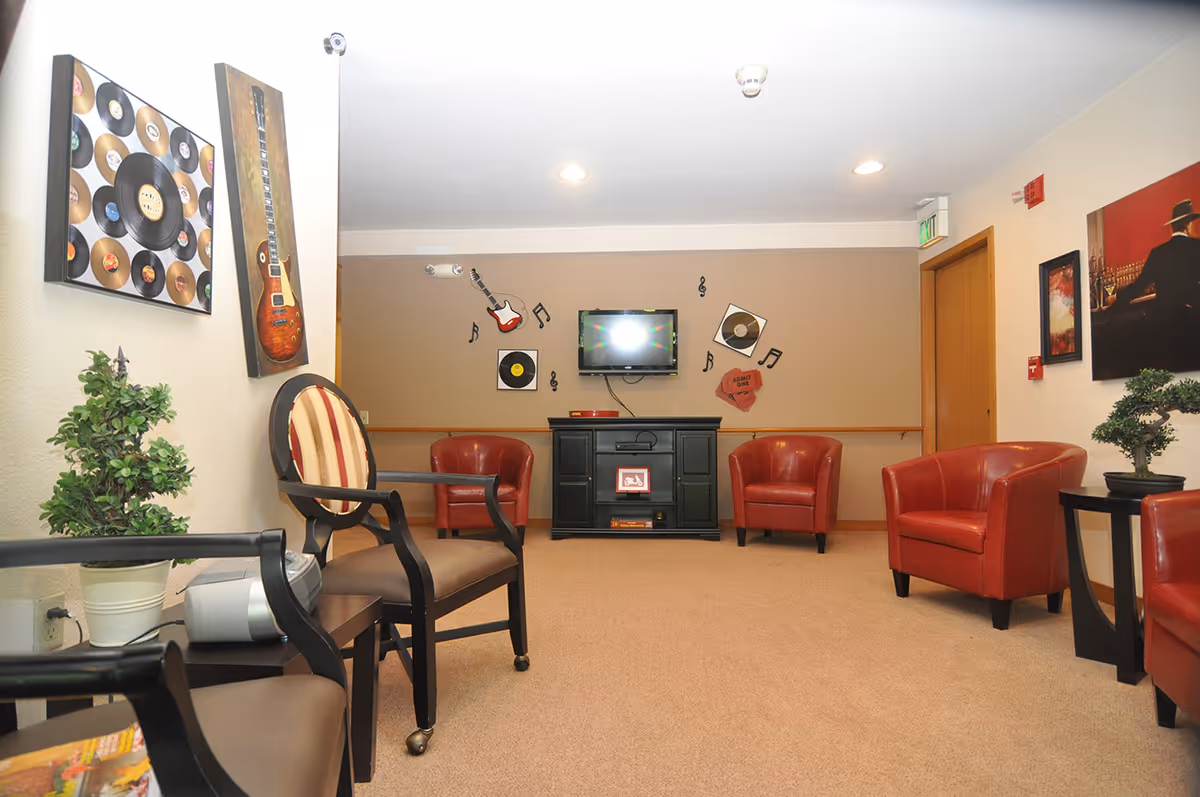 A cozy lounge area with red leather armchairs and a striped cushioned chair arranged around a black cabinet with a mounted TV above it. The walls are decorated with music-themed artwork including guitars, vinyl records, and musical notes. There are small potted plants on side tables and the room has beige carpeting and warm lighting.