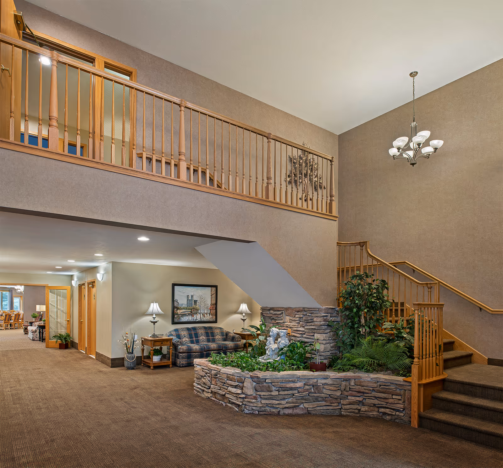 Spacious senior living facility lobby with seating, an indoor stone planter and a staircase under a chandelier.