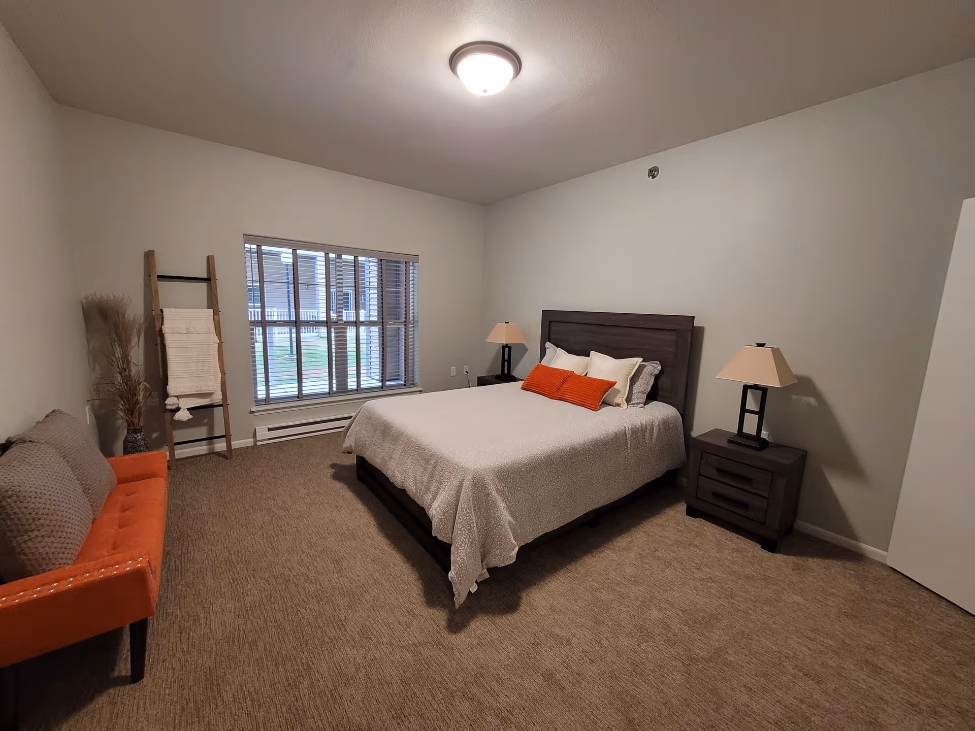 A bedroom with a large bed featuring white and orange pillows, two dark wooden nightstands with lamps, a window with blinds, an orange cushioned bench with a gray throw, and a decorative ladder with a white blanket leaning against the wall.