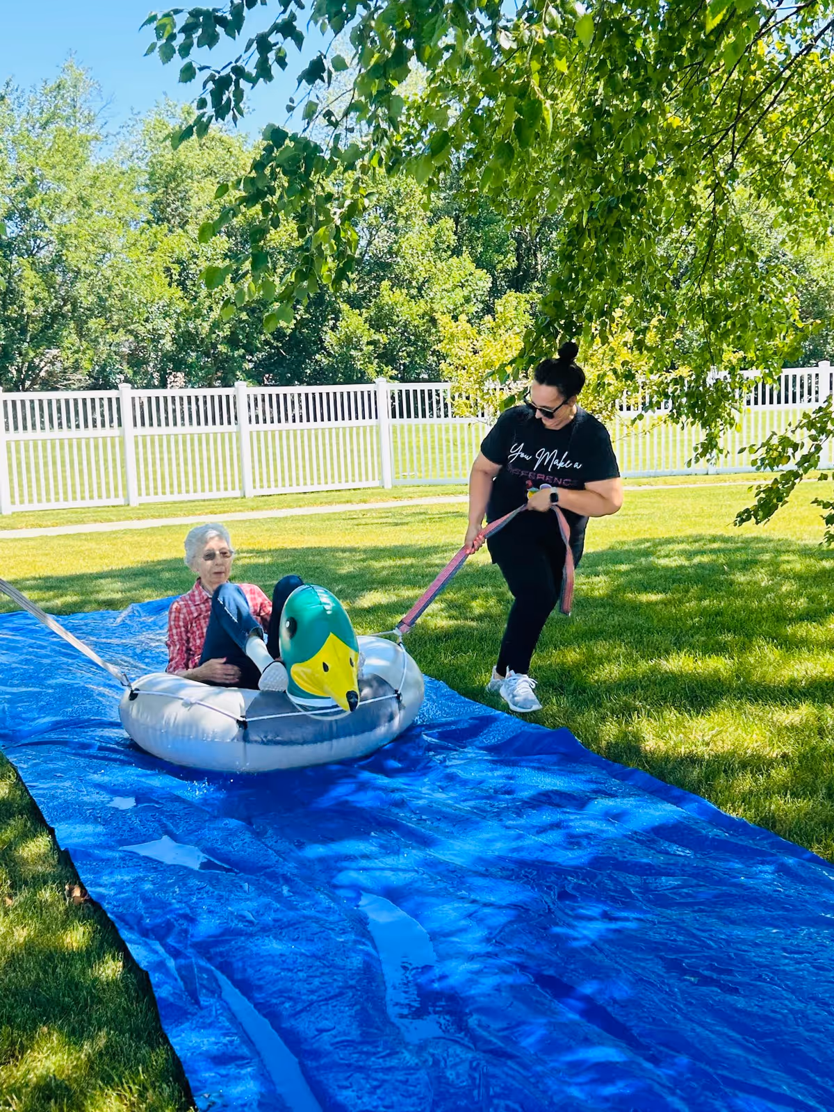 An elderly woman sitting in an inflatable duck-shaped raft on a blue tarp outdoors, being pulled by a younger woman holding a pink leash. They are in a grassy area with trees and a white fence in the background on a sunny day.