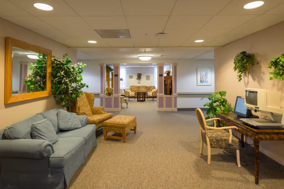 A cozy senior living facility common area with a blue couch and patterned armchair with ottoman on the left, a wooden table with a vintage computer and chair on the right, and a seating area with a sofa and chairs in the background. The walls are decorated with plants and framed artwork, and the space is well-lit with ceiling lights.