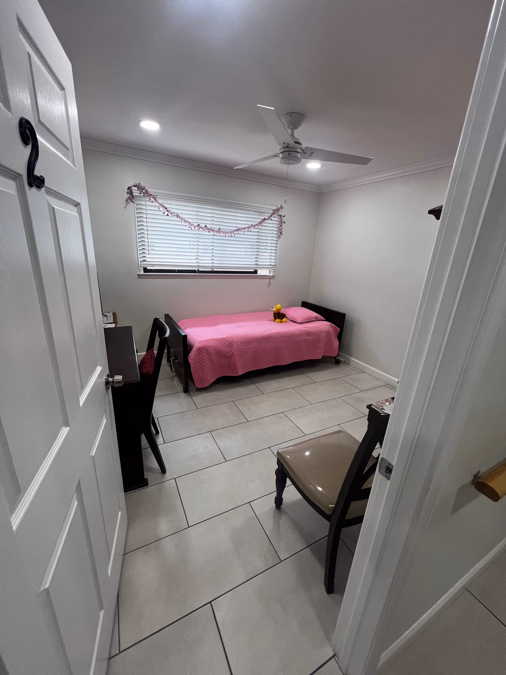 Small bedroom with a single bed covered in a pink quilt, a window with blinds and garland, a ceiling fan, tiled floor, and a couple of chairs.