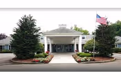 Front exterior view of Suffield House facility with a covered entrance, landscaped flower beds, two large evergreen trees, and an American flag on a flagpole to the right.