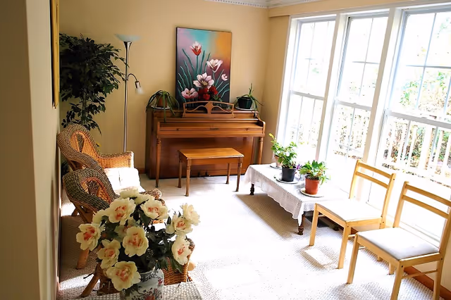 A bright living room with large windows letting in natural light. The room features a wooden piano with a matching bench, a floor lamp, wicker chairs with cushions, a small table with potted plants, and a vase of flowers in the foreground.