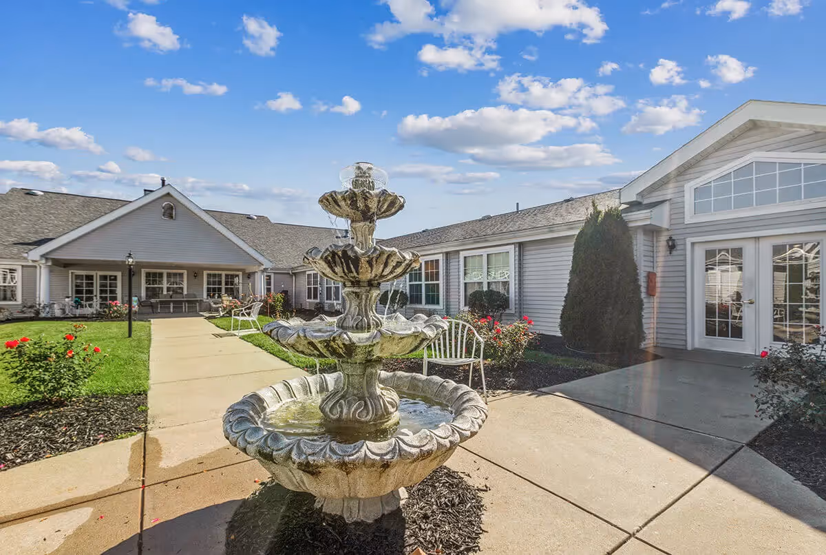 Outdoor courtyard area of The Addison of Garden Way Place featuring a three-tier stone water fountain in the center, surrounded by a concrete walkway, green grass, rose bushes, white benches, and the exterior of the facility building under a blue sky with scattered clouds.