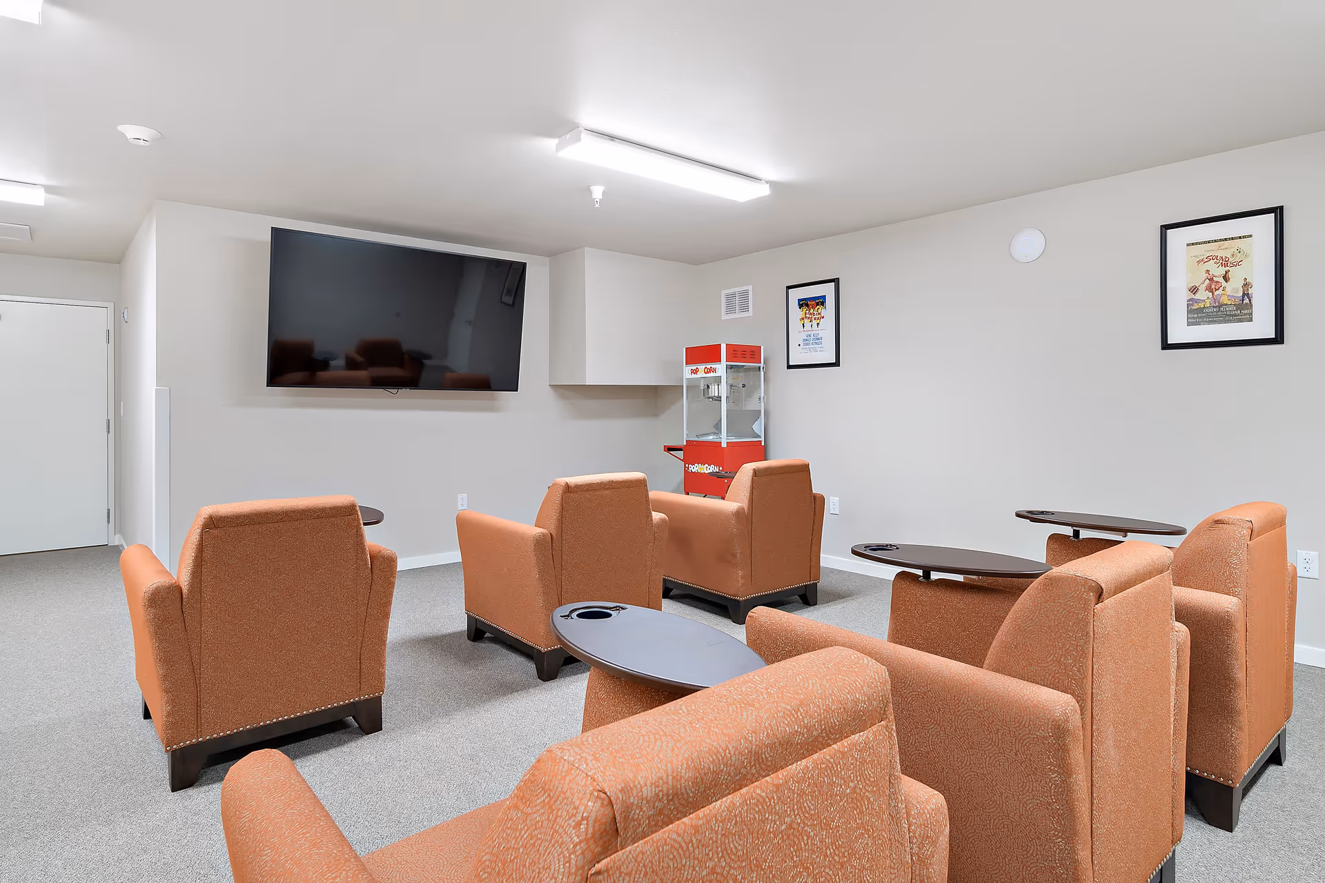 A small screening room with rows of orange upholstered chairs facing a wall-mounted TV and a popcorn machine.