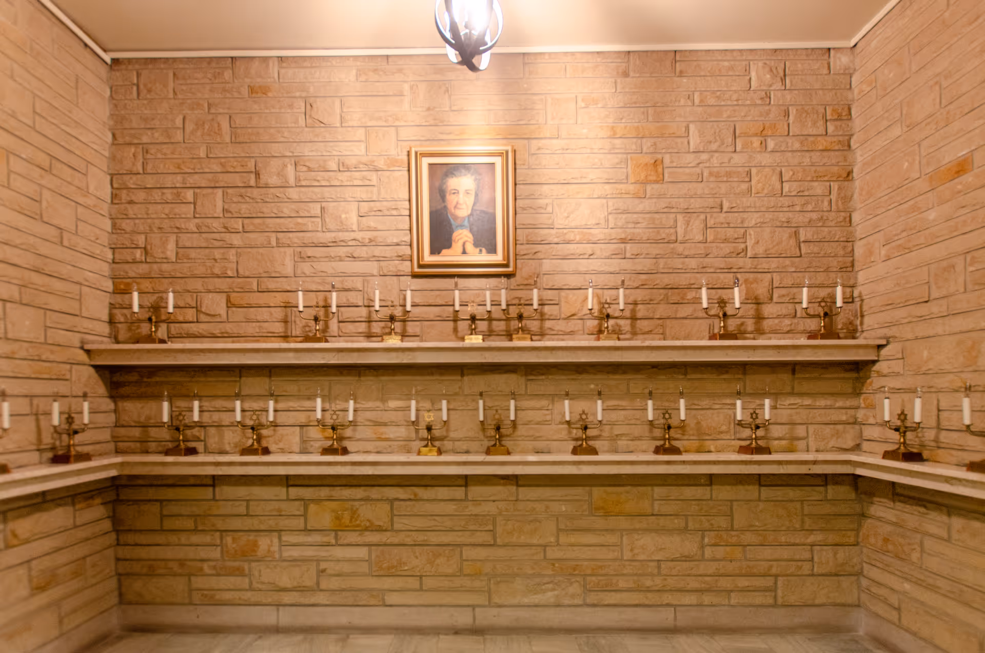 A stone-walled interior room with shelves of brass candelabras and a framed portrait on the wall.