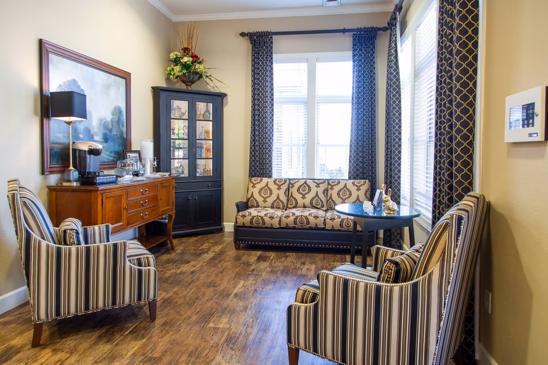 A cozy sitting area in an assisted living facility with two striped armchairs, a patterned sofa, a small round table, a wooden sideboard with a coffee maker and lamp, a black cabinet with glass doors, and large windows with patterned curtains letting in natural light.