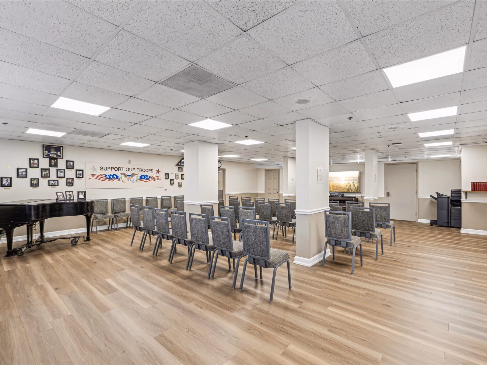 A large room with rows of chairs arranged facing a wall with a 'Support Our Troops' banner and framed photos. There is a black grand piano on the left side and a printer or copier on the right side near a door. The room has a drop ceiling with fluorescent lights and wood-patterned flooring.