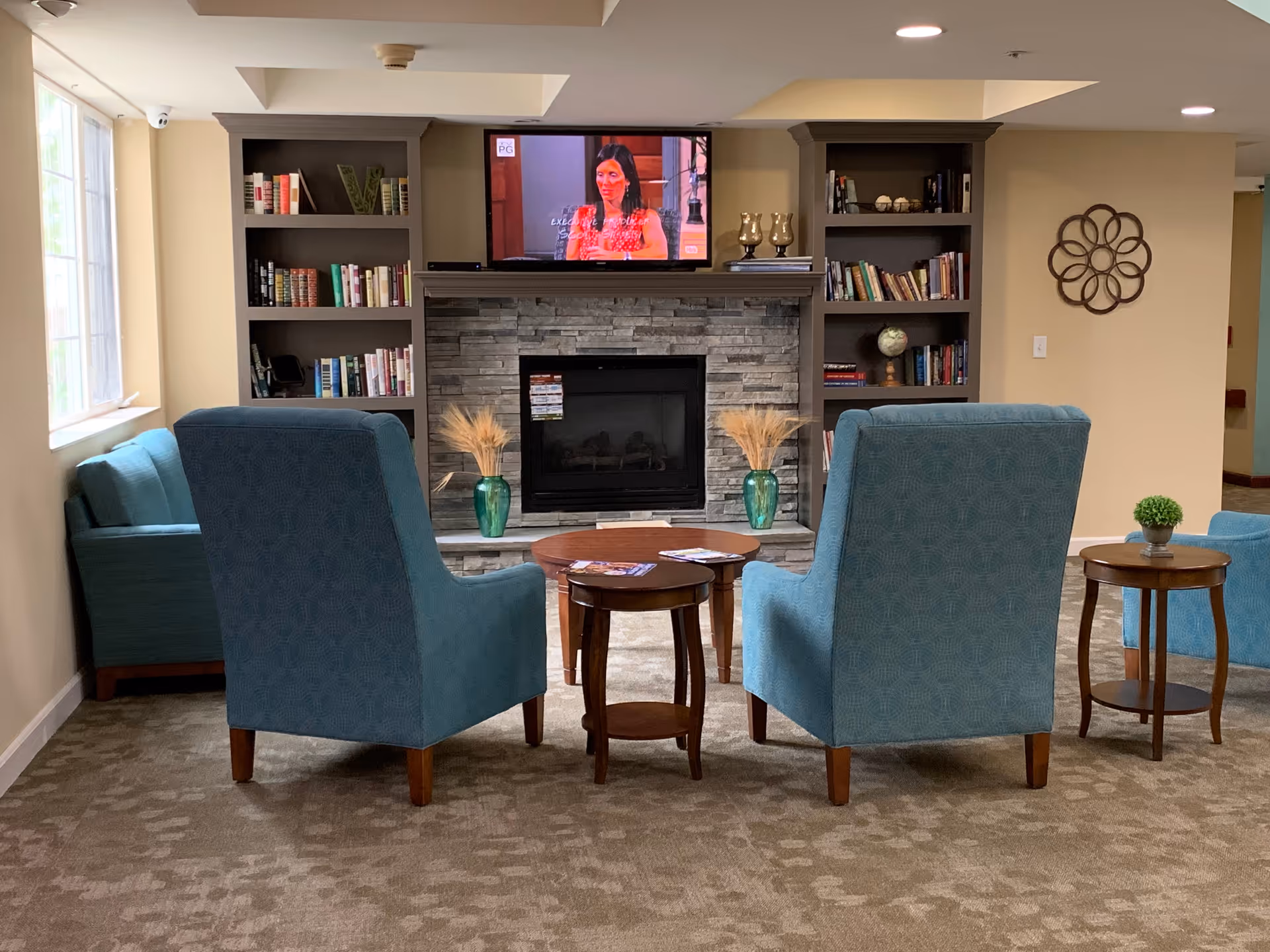 A cozy living room area with two blue armchairs and a blue sofa arranged around a wooden coffee table and side tables. A stone fireplace with two green vases holding dried wheat stalks is centered on the back wall, flanked by built-in bookshelves filled with books and decorative items. Above the fireplace, a television is mounted on the wall showing a woman speaking. The room has beige walls, a patterned carpet, and a window letting in natural light on the left side.