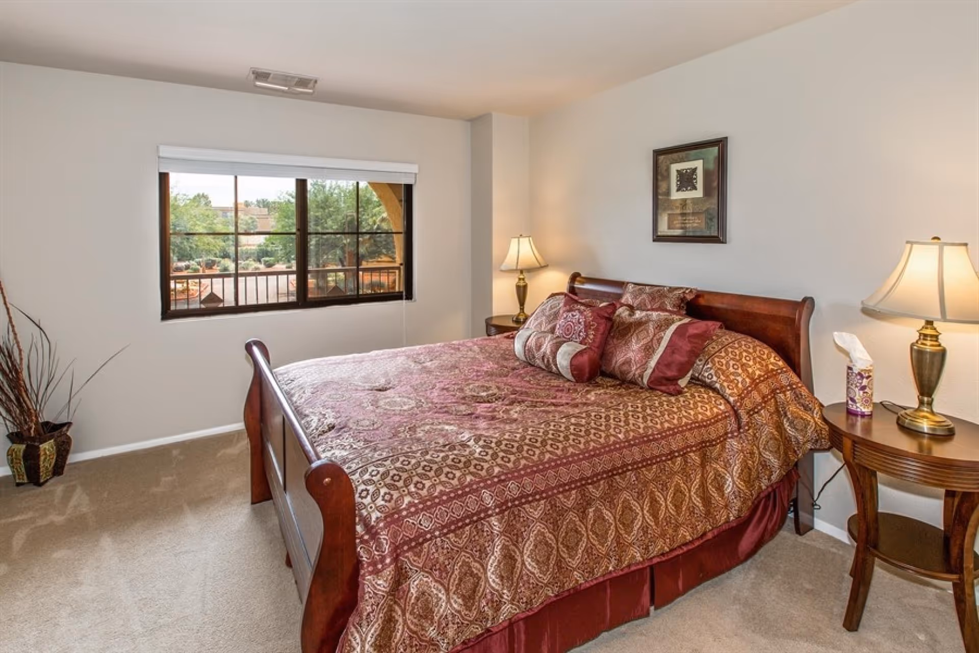 A bedroom with a large wooden bed covered in a patterned red and gold bedspread with multiple decorative pillows. There are two wooden nightstands on either side of the bed, each with a brass table lamp. A framed picture hangs on the wall above the bed. A window with a white blind shows a view of trees and outdoor structures. A decorative vase with tall dried branches is placed on the carpeted floor near the window.