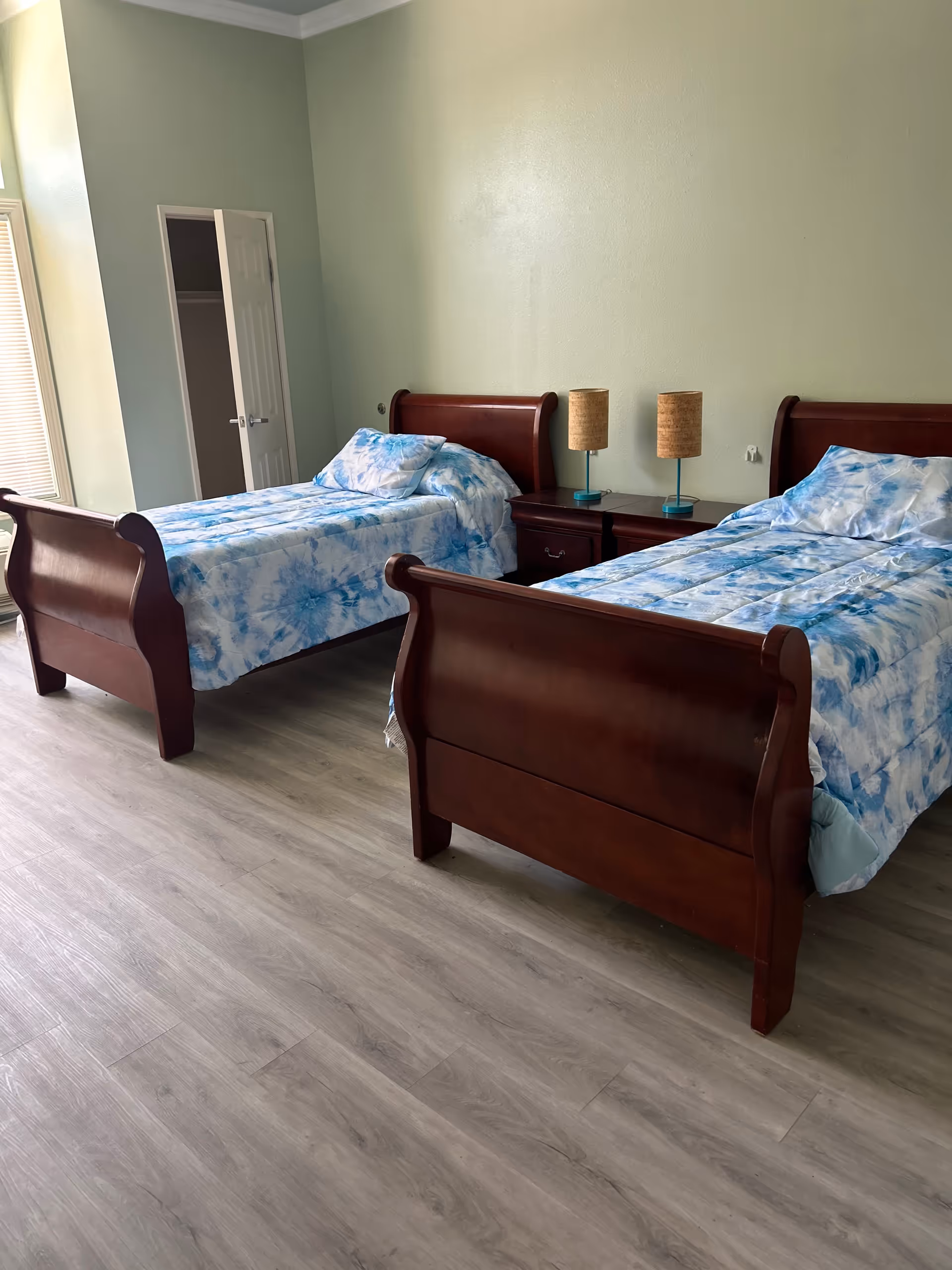 Room with two wooden twin beds with blue-patterned bedding, a shared nightstand with lamps, and a light green wall.