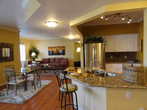 Open concept interior of a senior living facility showing a kitchen with granite countertops, a sink, and bar stools in the foreground. In the background, there is a dining area with a glass table and chairs on a rug, and a living room area with a red sofa, a coffee table, a floor lamp, and a colorful painting on the wall. The walls are painted yellow and the floor is wooden.
