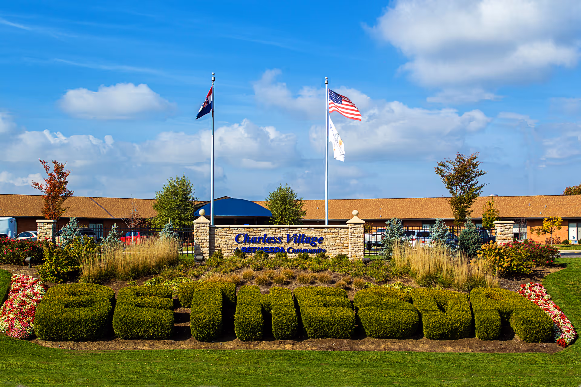 Outdoor view of a senior living facility with a large neatly trimmed hedge spelling out the word 'SENIORS' in front of a stone sign that reads 'Charles Village Retirement Community'. Two flagpoles with flags are behind the sign, and the building with a brown roof is visible in the background under a partly cloudy blue sky.