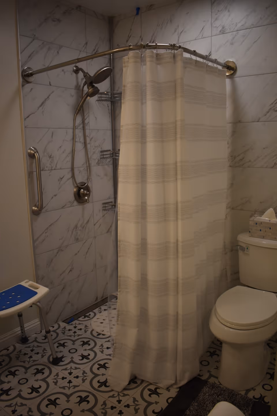 Bathroom with a shower behind a curtain, grab bars, a shower stool, and a toilet on patterned tile flooring.