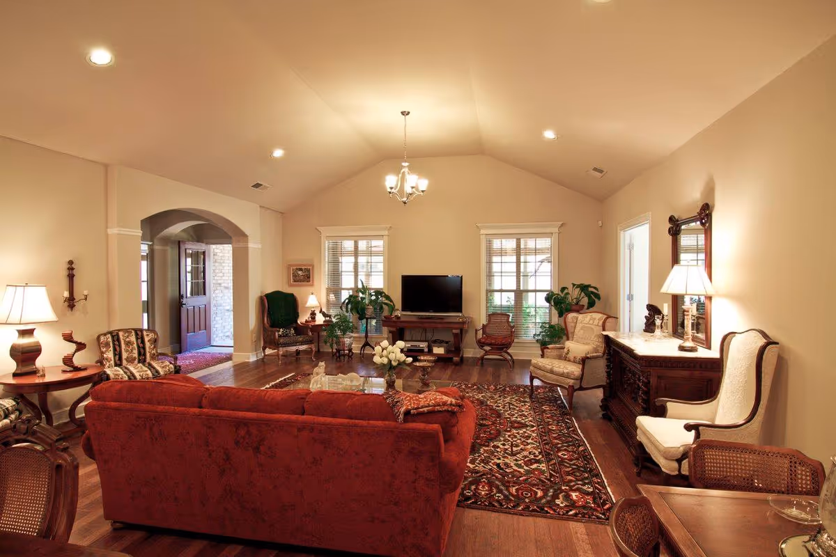 A warmly lit living room with a red sofa, patterned armchairs, a wooden coffee table with a vase of white flowers, and a patterned rug. The room features hardwood floors, two windows with blinds, a TV on a wooden stand, several plants, and various lamps providing soft lighting. There is an arched doorway leading to another area and a wooden sideboard with a mirror and lamp on the right side.