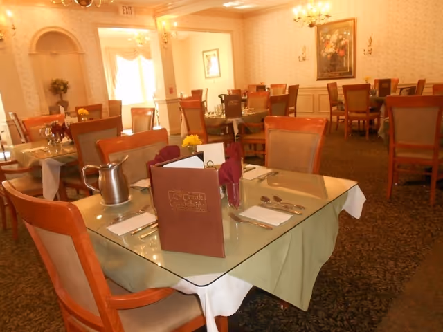 A dining room with multiple tables set for meals. Each table has a glass top, cloth napkins, silverware, and a menu. The room has patterned wallpaper, carpeted floors, chandeliers, and framed artwork on the walls. There is a pitcher on one table and floral centerpieces on several tables.