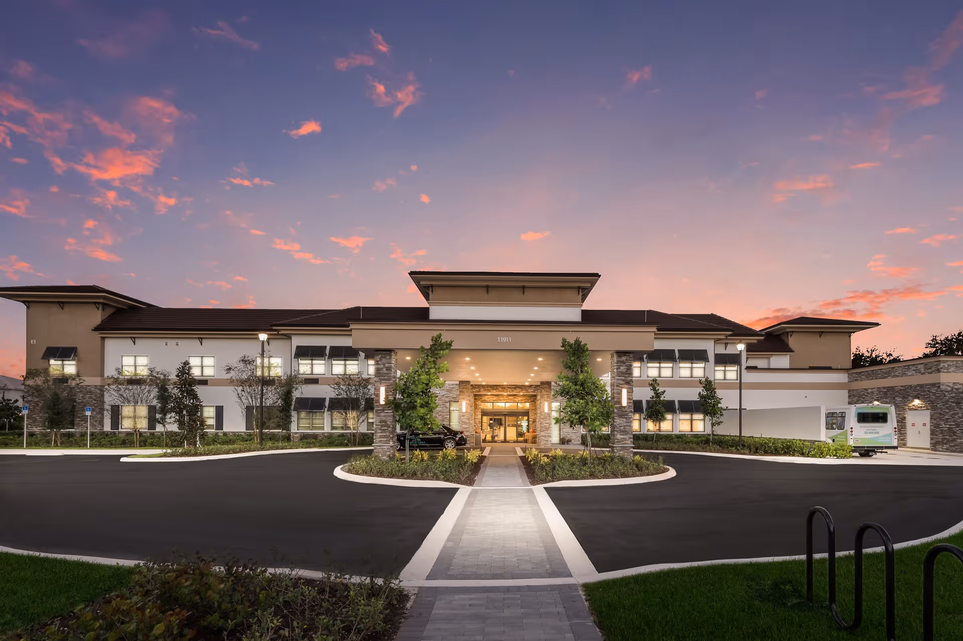 Front exterior view of Bella Mar Royal Palm Beach facility at dusk with a paved driveway, landscaped greenery, and a clear sky with pink clouds.