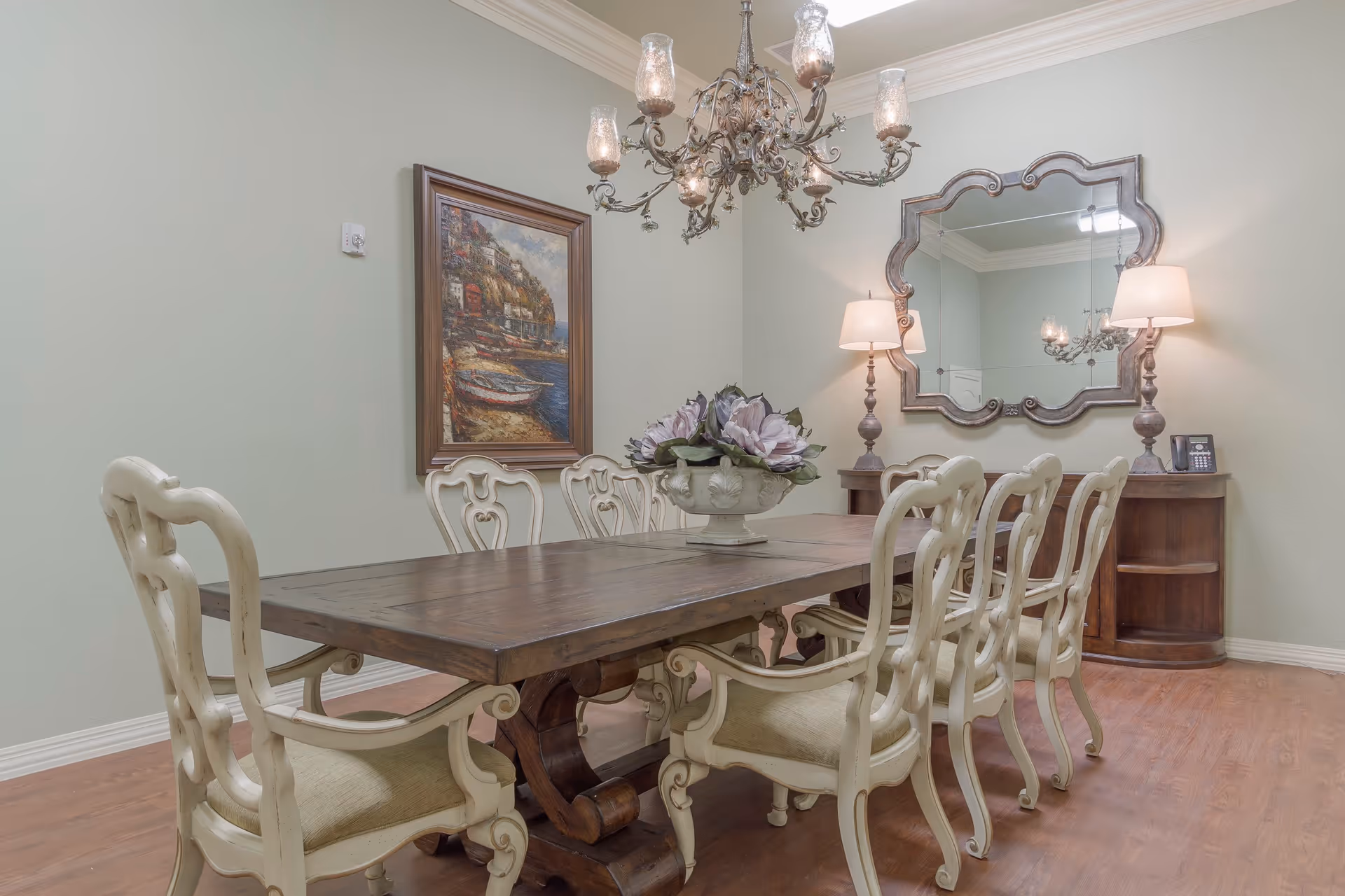 A dining room with a large wooden table surrounded by eight cream-colored upholstered chairs. A decorative chandelier hangs above the table. On the table is a floral centerpiece. The room has light green walls, a framed painting on one wall, and a large ornate mirror above a wooden sideboard with two lamps and a phone.
