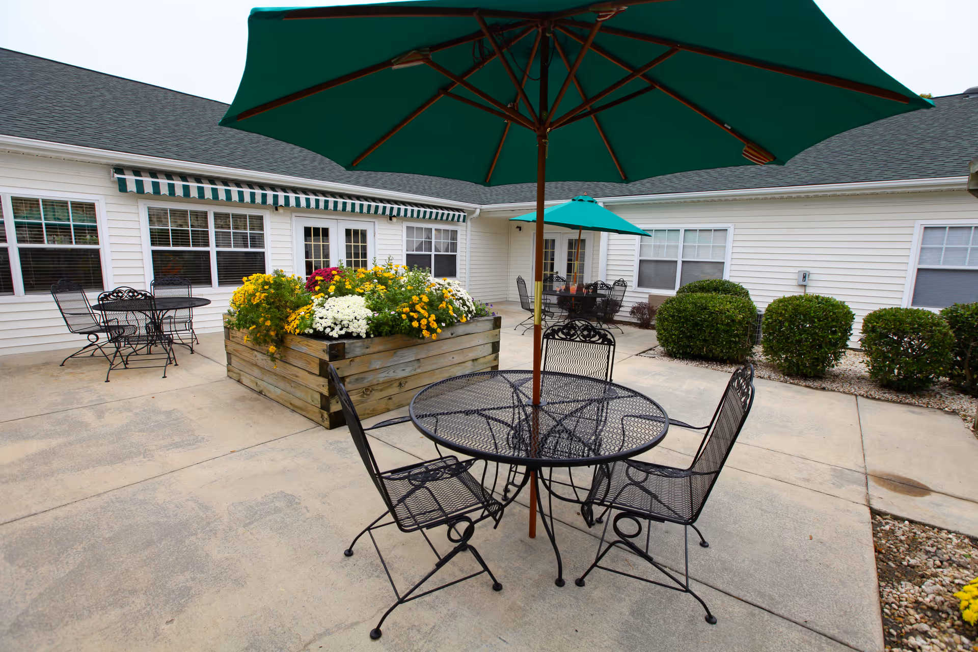 Outdoor patio area with black metal tables and chairs, each table shaded by a green umbrella. There is a large wooden planter box filled with colorful flowers in the center. The patio is surrounded by a white building with multiple windows and green bushes along the edges.