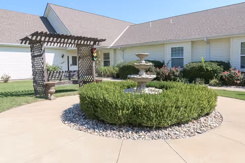 Outdoor courtyard area of a senior living facility with a circular hedge surrounding a three-tiered stone fountain, a wooden pergola with a bench swing, and a building with beige siding and multiple windows in the background under a clear blue sky.