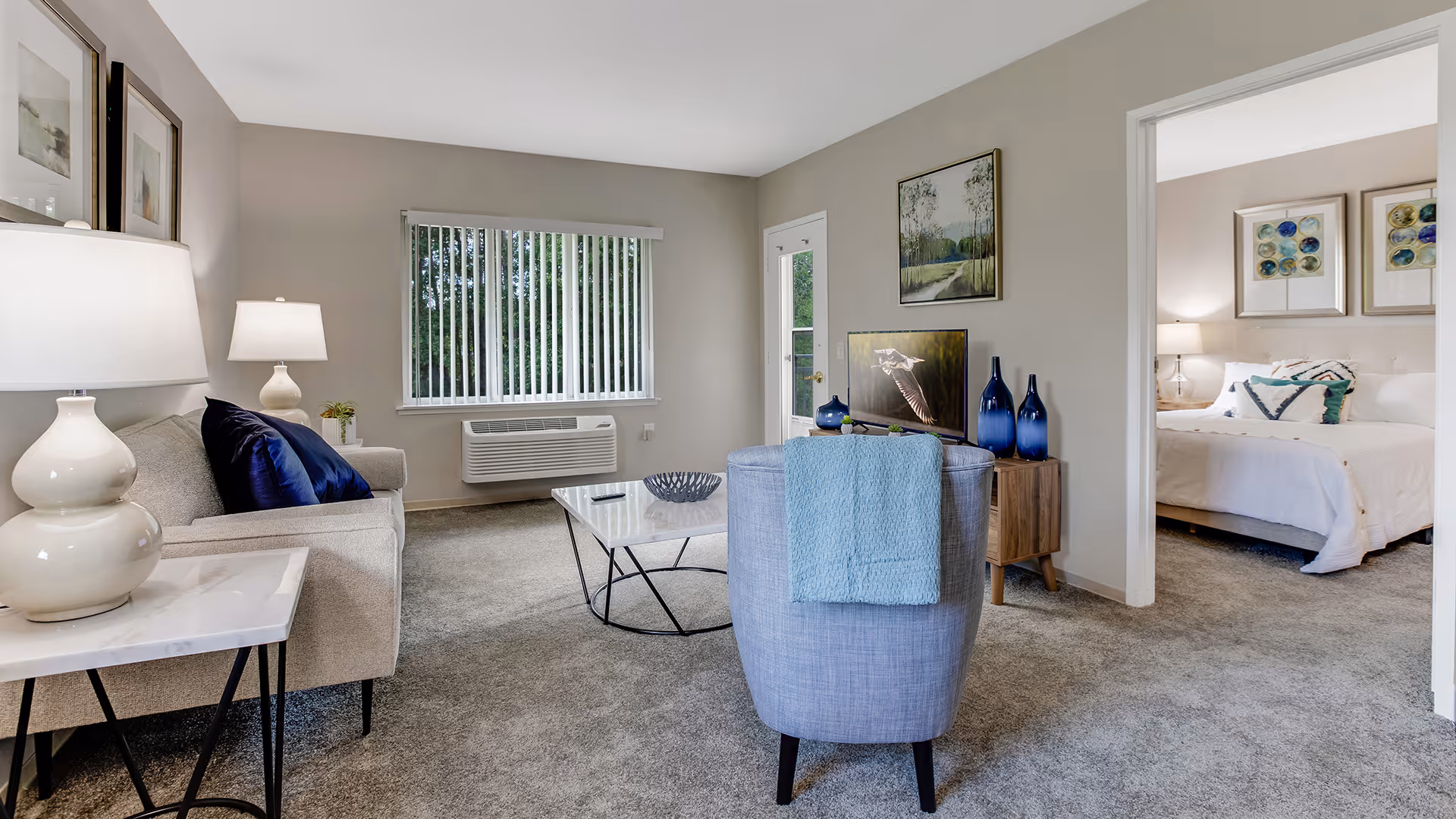 A cozy living room with a beige sofa adorned with dark blue pillows, a light blue armchair with a blue throw draped over it, and a glass coffee table. There is a window with vertical blinds and an air conditioning unit below it. A small wooden cabinet holds decorative blue vases and a TV. The room leads into a bedroom with a neatly made bed, bedside lamps, and framed artwork on the walls.