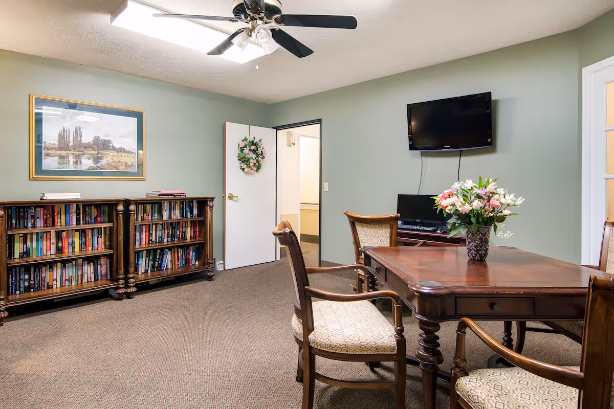 A cozy room with green walls featuring a wooden table with four upholstered chairs around it. A vase with pink and white flowers is placed on the table. On one wall, there is a flat-screen TV mounted above a small desk with a computer. A bookshelf filled with books is against another wall beneath a framed landscape painting. The room has a ceiling fan with lights and an open door decorated with a floral wreath.