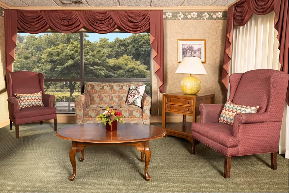 A cozy living room area with two maroon upholstered armchairs, a patterned loveseat with decorative pillows, a wooden coffee table with a small floral arrangement, and a wooden side table with a yellow lamp. Large windows with maroon drapes offer a view of green trees outside.