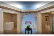 Interior view of a senior living facility bathroom area with two separate bathing spaces on either side of an arched doorway. The left side features a walk-in bathtub, and the right side has a bathtub with a decorative niche above it. In the center, there is a small round table with a potted orchid plant and a small statue, under soft ceiling lighting.