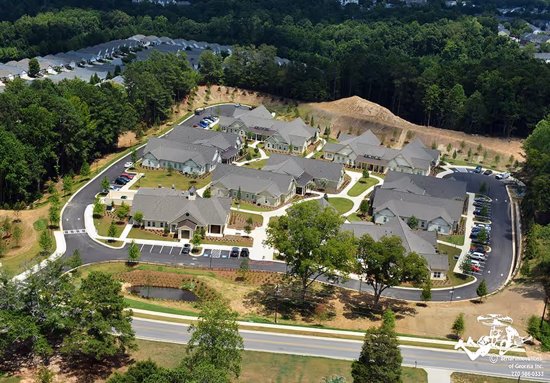 Aerial view of Camellia Place an Oaks Senior Living Community showing multiple single-story buildings with gray roofs arranged around paved roads and walkways, surrounded by trees and greenery.