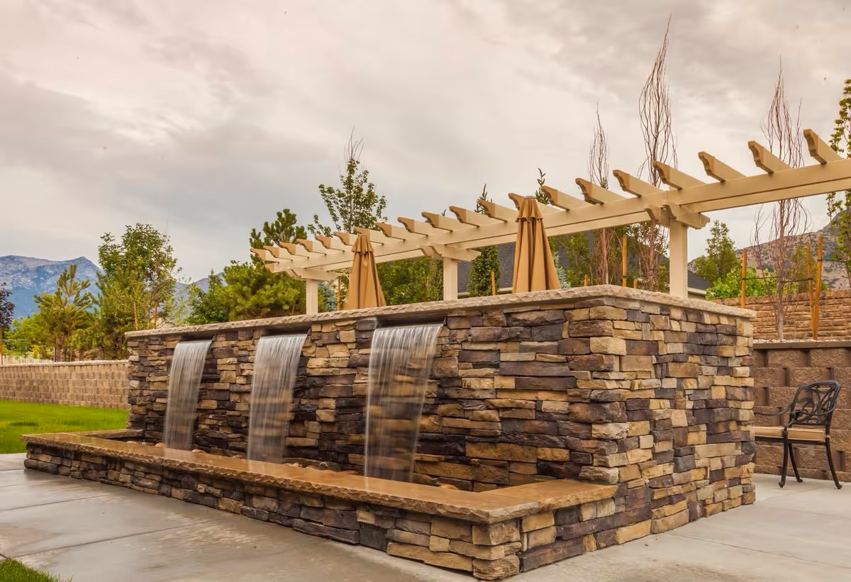 Outdoor stone water feature with three cascading waterfalls, surrounded by greenery and trees, with a pergola structure and umbrellas in the background under a cloudy sky.