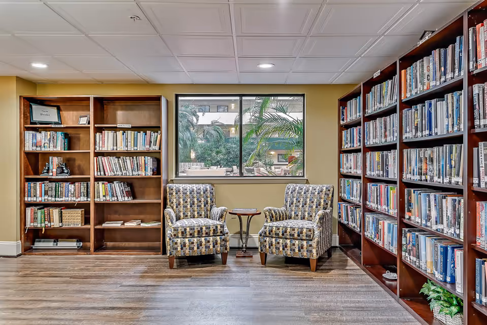 A cozy library room with wooden bookshelves filled with books on the left and right sides. Two patterned armchairs are placed in front of a window that shows an outdoor courtyard with greenery. A small round table is positioned between the chairs.
