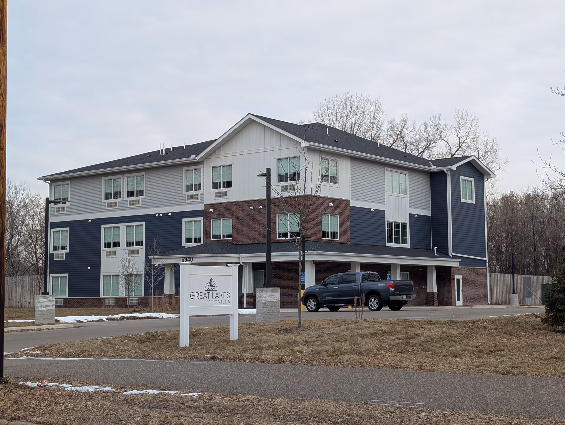 Exterior view of a three-story senior living facility building with a mix of brick and siding in gray and blue tones. A sign in front reads 'Great Lakes Villa'. There is a black pickup truck parked near the entrance, and leafless trees and a wooden fence surround the property.