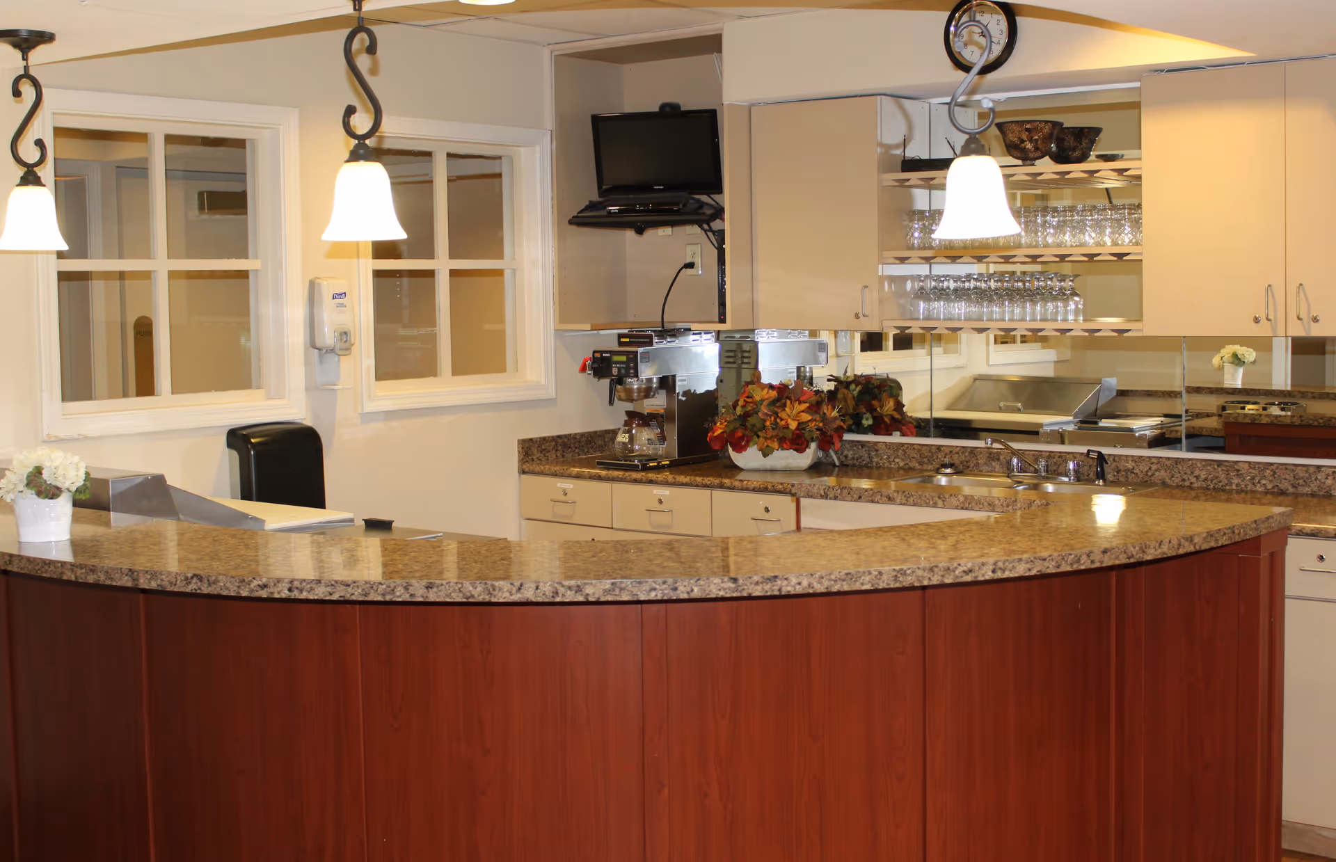 Interior view of a reception or service counter area with a curved granite countertop, wooden paneling, two hanging pendant lights, a coffee maker, a small TV mounted on the wall, and shelves with glassware. There are two windows on the wall behind the counter and a clock above the cabinets.