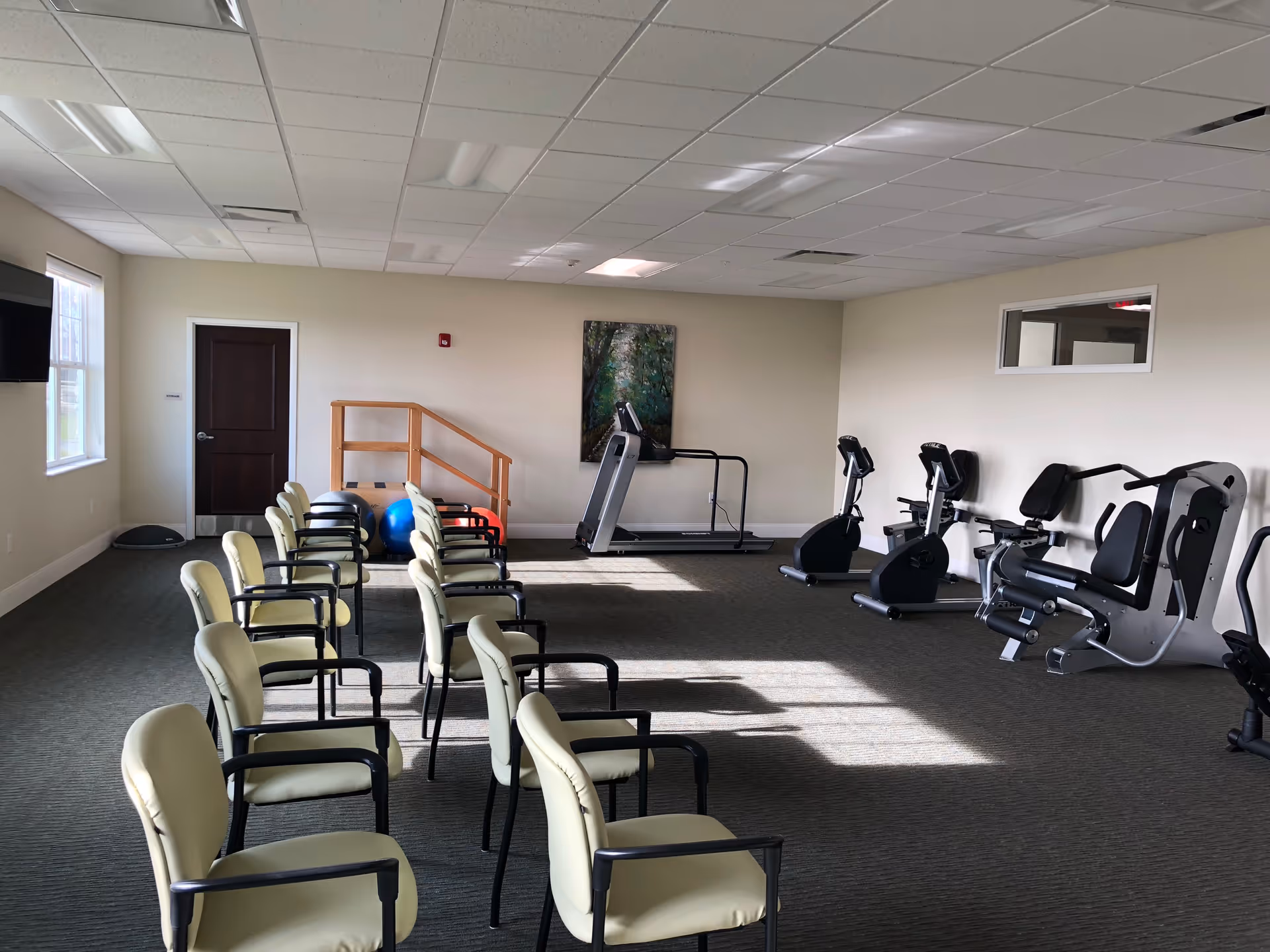 A bright interior fitness room with rows of chairs and exercise equipment like a treadmill, recumbent bikes, and strength machines.
