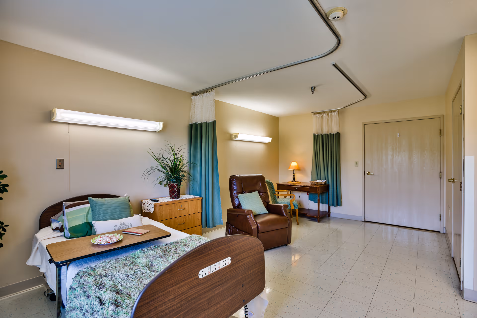 A nursing center bedroom with a hospital bed covered in a green and white blanket, a wooden bedside table with a plant, a brown recliner chair with a green pillow, a small desk with a lamp and chair, and curtains dividing the room. The floor is tiled, and the walls are painted beige.