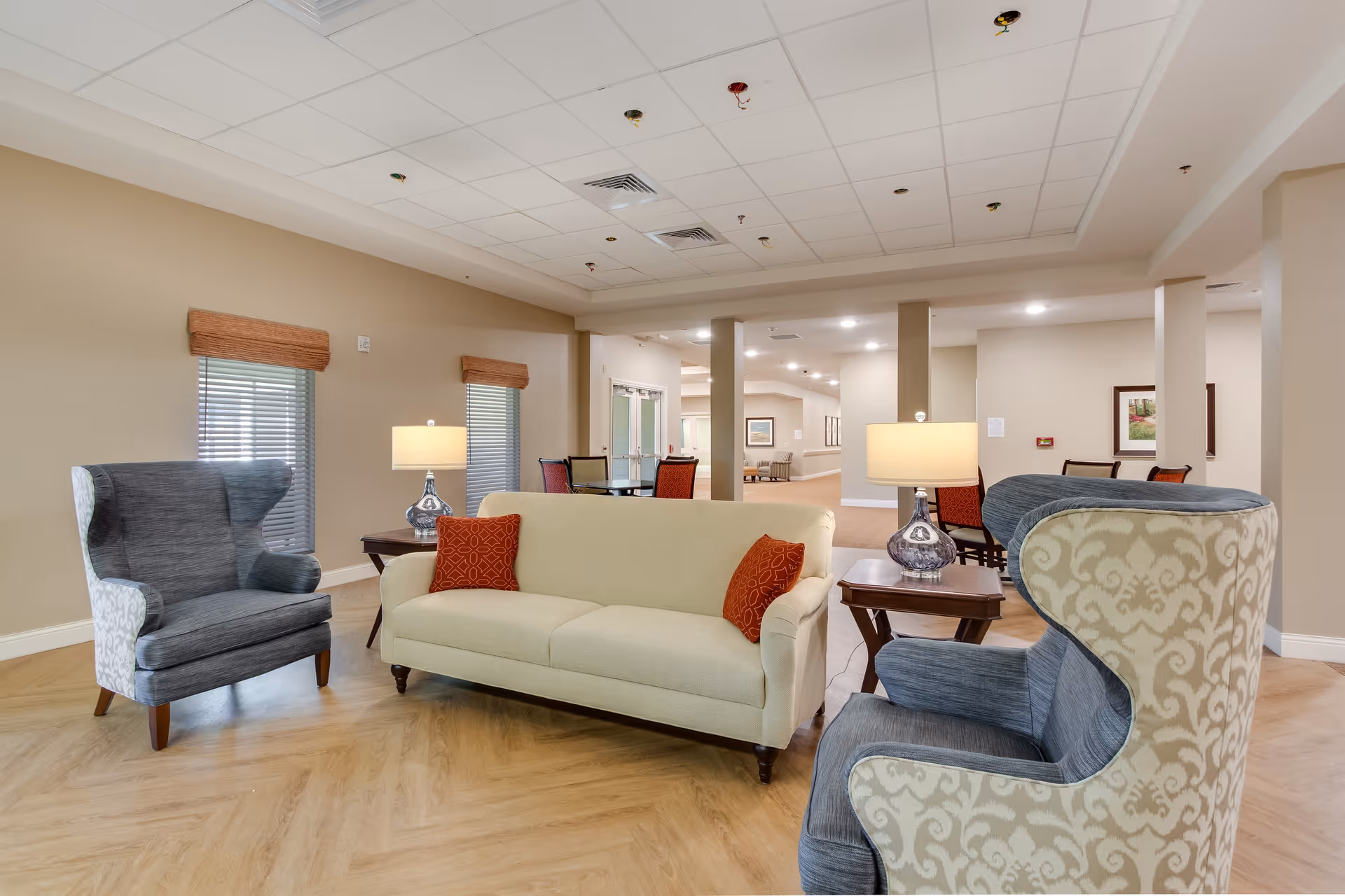 A spacious and well-lit common area in an assisted living facility featuring a beige sofa with two red patterned cushions, two wingback armchairs with a blue and white pattern, two side tables with lamps, and a dining area with chairs in the background. The room has light-colored walls, wood-patterned flooring, and windows with wooden blinds.
