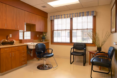 A well-lit room with wooden cabinets and a countertop on the left side, featuring a white sink. There are three black chairs arranged in the room, one near the sink and two against the wall under a large window with blinds and a valance. A small table with a plant is positioned near the chairs on the right side. The walls are painted beige and the floor is light-colored.