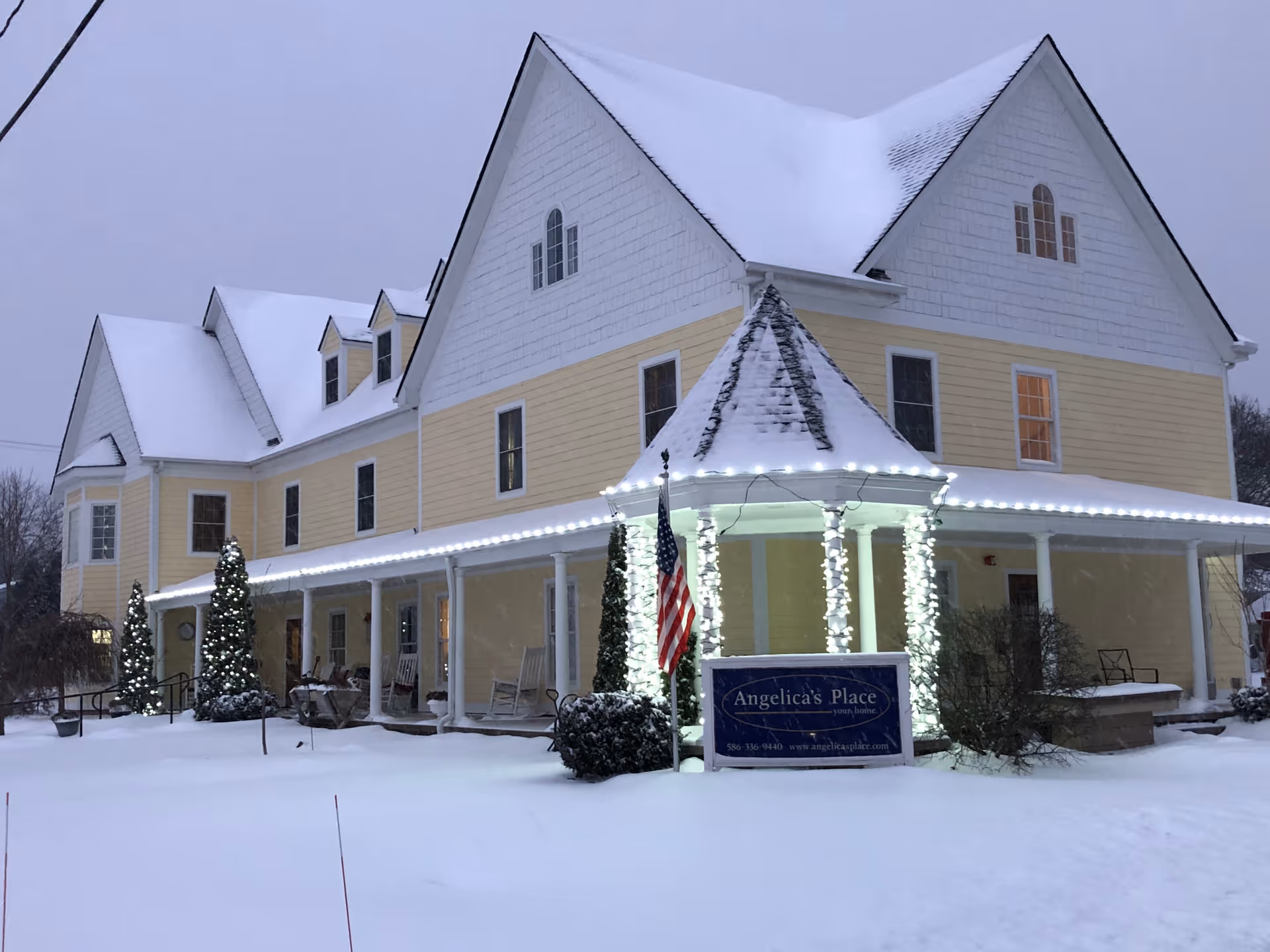 A large two-story yellow and white building covered in snow with a steep roof also covered in snow. The building has a wrap-around porch decorated with white string lights and an American flag. There are small decorated evergreen trees with lights near the entrance. A sign in front reads 'Angelica's Place'. The scene is set during winter with snow on the ground and a gray sky.