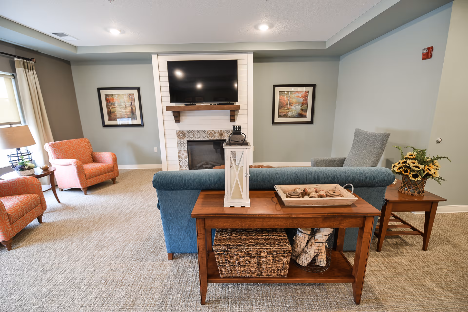 Comfortable common living area with a blue sofa facing a wall-mounted TV above a fireplace, flanked by armchairs and side tables.