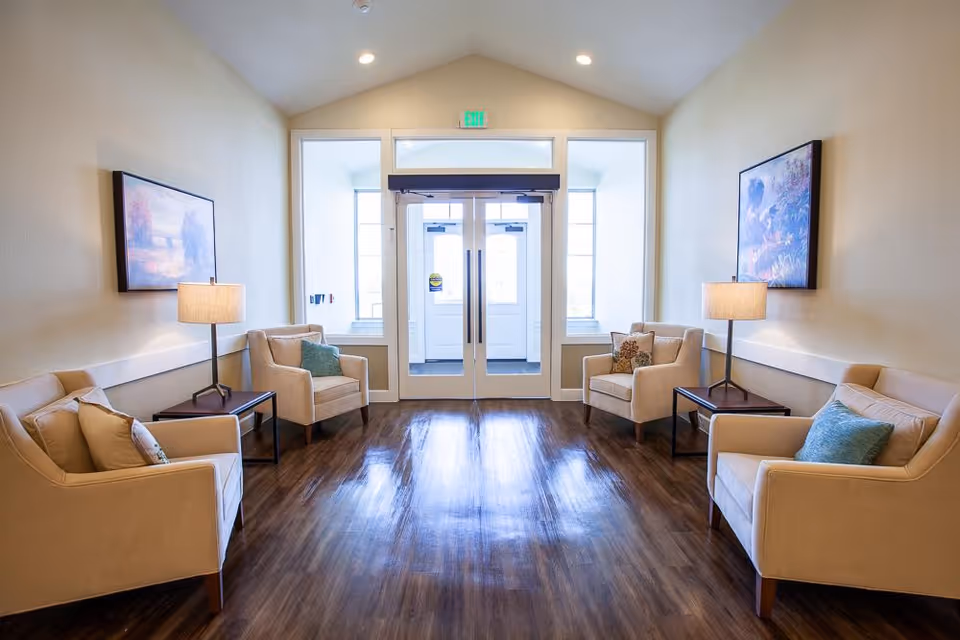 A bright and clean seating area with four beige armchairs arranged in pairs on either side of a wooden floor. Each pair of chairs is separated by a small dark wooden side table with a lamp. Two framed paintings hang on the light-colored walls above the chairs. At the far end, there is a set of glass double doors with windows on either side, allowing natural light to fill the space.