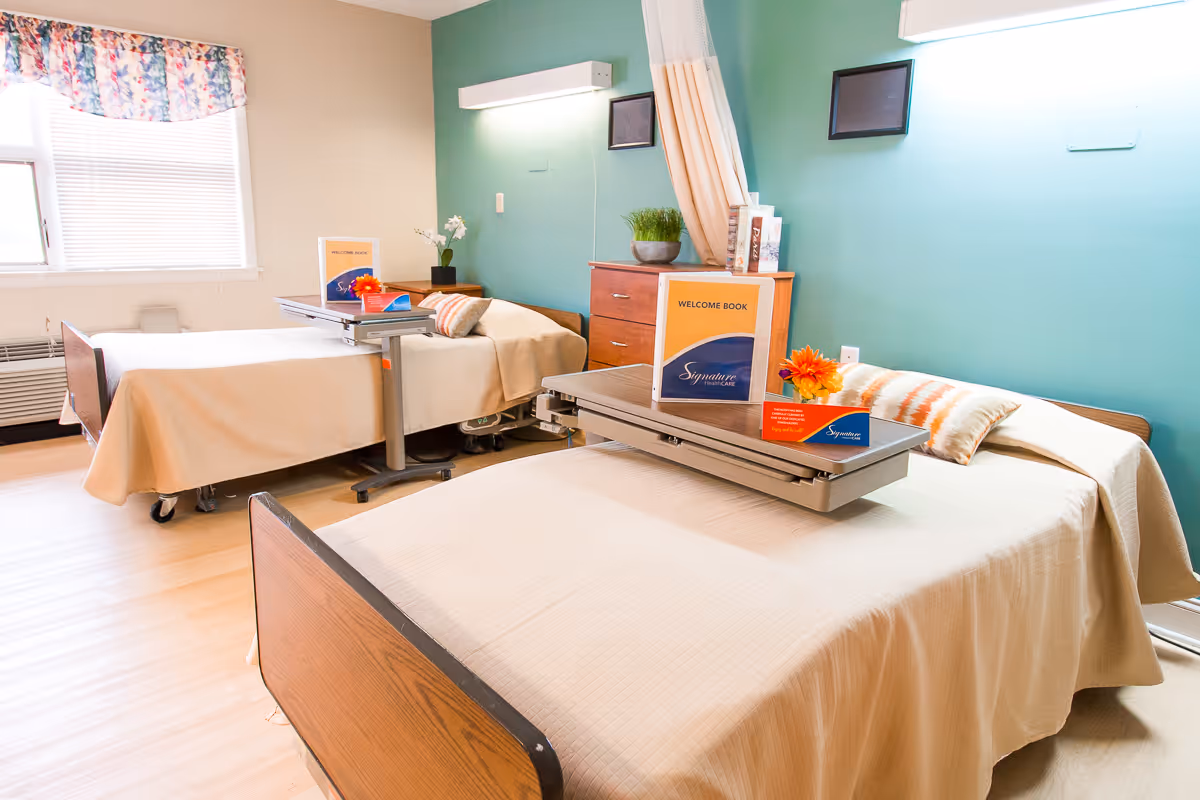 Two hospital-style beds in a bright, tidy patient room with a teal accent wall and overbed tables holding welcome brochures.