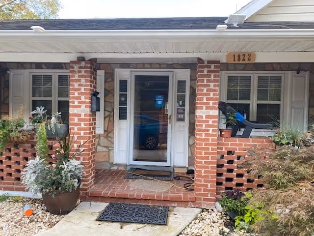 Front entrance of a single-story brick building with a white door and windows on either side. The entrance is framed by two brick pillars and has potted plants and greenery around the porch area. The building number 1822 is displayed above the door.