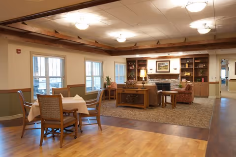 A warm communal dining and living room with wooden floors, tables and chairs, a seating area around a fireplace, and built-in shelving.