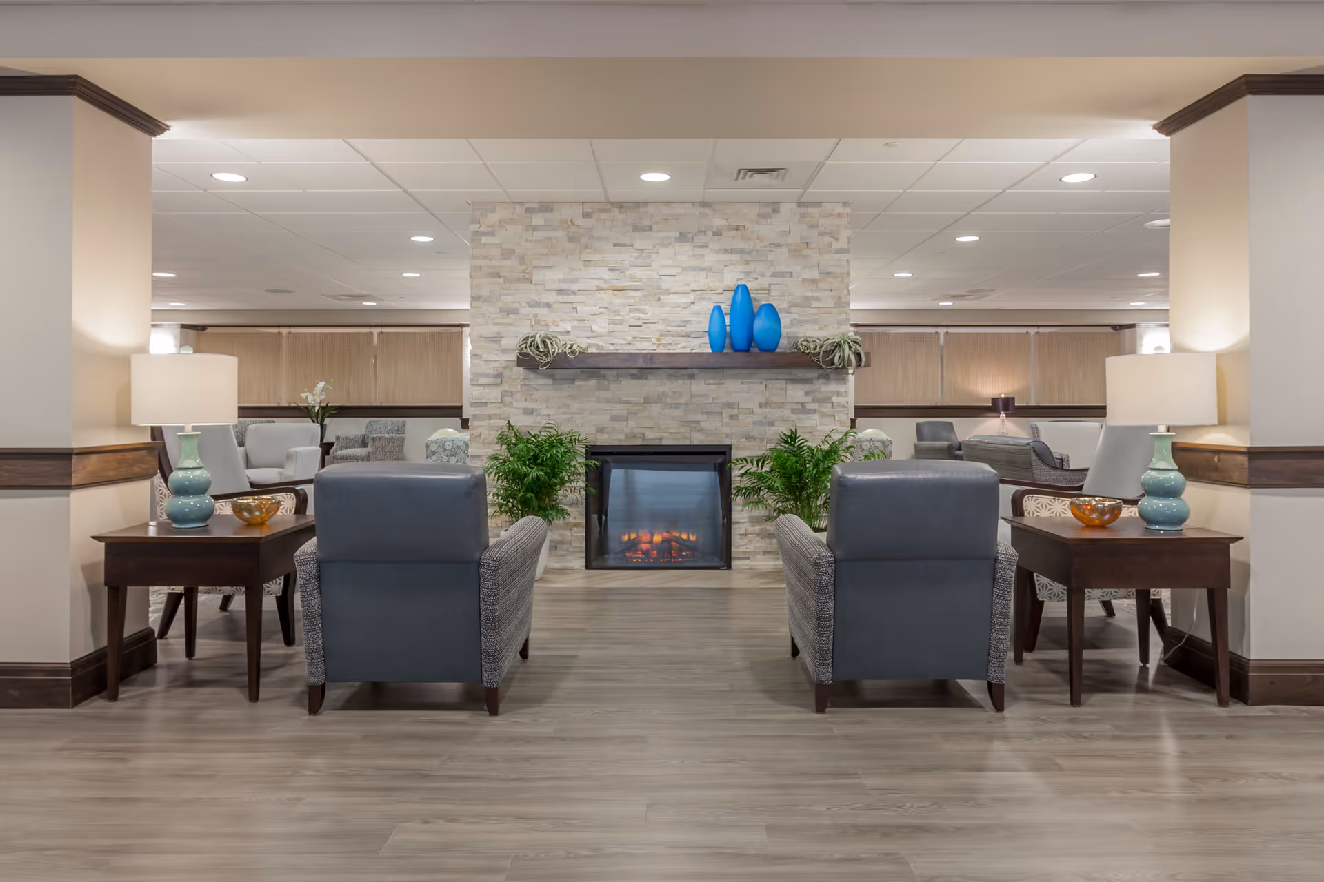 A furnished lounge with two armchairs facing a stone fireplace, side tables with lamps, and decorative plants and vases.