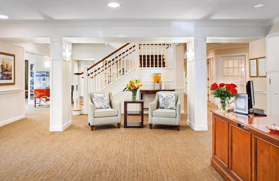 A bright and welcoming interior space with beige carpet and white walls. Two light gray armchairs with patterned cushions are positioned around a small dark wooden table holding a vase of flowers. Behind the chairs is a staircase with white railings and wooden handrails. To the right, there is a wooden reception desk with a computer and a vase of red roses. The left side shows a popcorn machine and a framed painting on the wall.