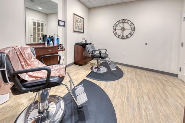 Interior view of a salon area in a senior living facility featuring two black salon chairs with footrests on black mats, a wooden cabinet with hair care products and a large mirror, a decorative wall clock, and light-colored wood flooring.