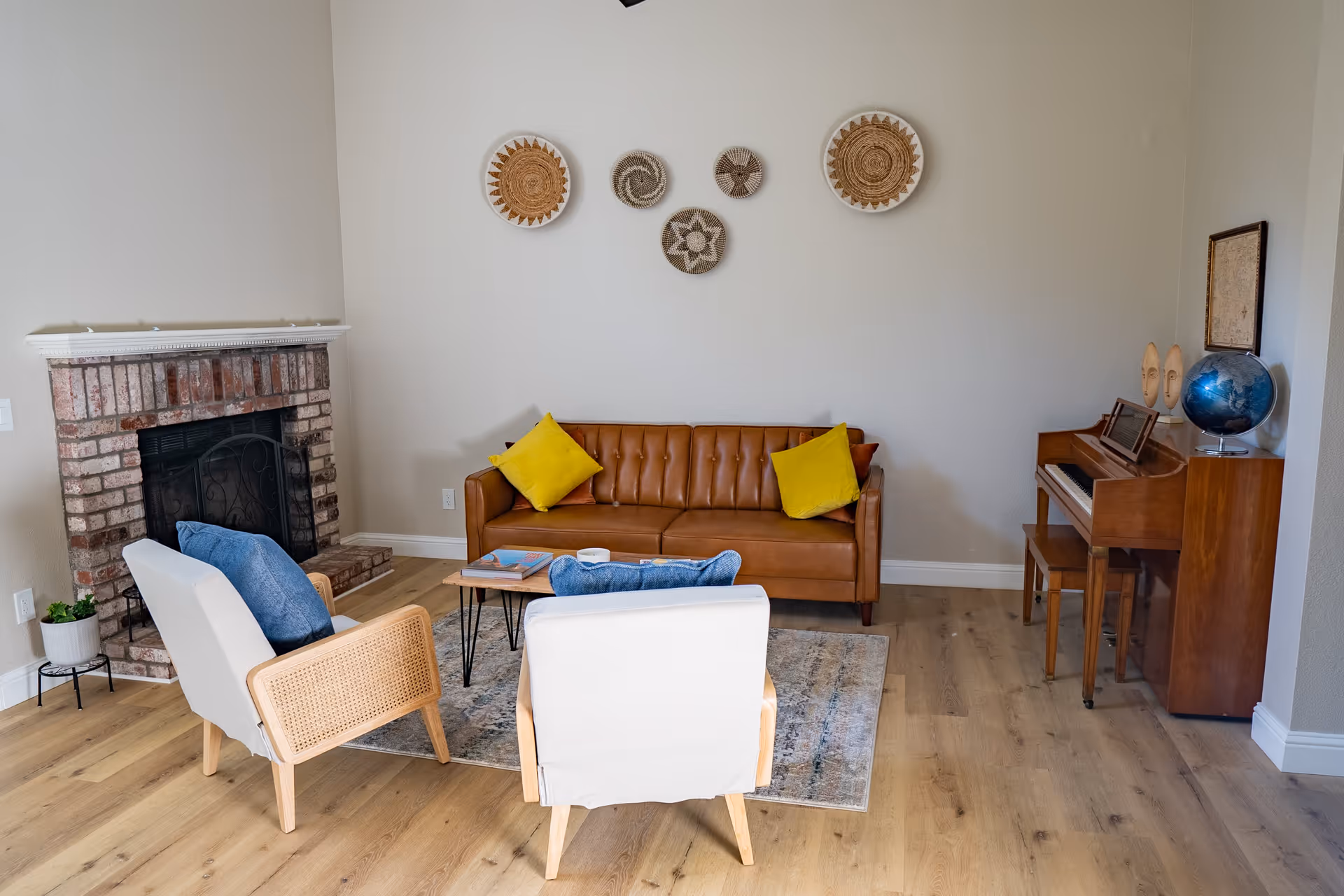 A cozy living room with a brown leather sofa adorned with yellow pillows, two white armchairs with blue cushions, a small wooden coffee table, a brick fireplace, a wooden piano with a globe and decorative masks on top, and woven wall art hanging on a beige wall.