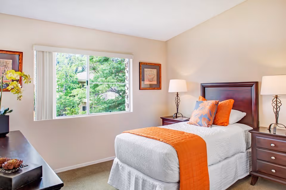 A bright and clean bedroom with a single bed dressed in white linens and an orange quilted throw blanket. The bed has two decorative pillows, one orange and one with a leaf pattern. There are two matching dark wood nightstands on either side of the bed, each with a lamp. A large window with vertical blinds shows green trees outside. The walls are light-colored with two framed pictures hanging.